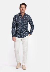 Chemise à manches longues, bleu marine avec un motif floral, fermeture boutonnée à l'avant, associée à un pantalon blanc et des chaussures à enfiler beiges.