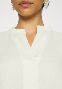 Haut en lin blanc avec un col en V et un petit col slim ; le tissu présente une texture légèrement translucide. Boucles d'oreilles goutte en perle.