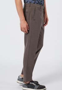 Brown tailored trousers with a subtle check pattern, featuring side pockets and a logo, paired with black and gray sneakers.