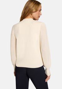Femme aux longs cheveux blonds portant un pull crème texturé à manches longues et un pantalon noir, vue de dos sur un fond blanc.