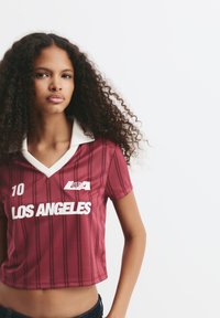 Ung kvinde med krøllet hår iført en maroon stribet sportsjersey med teksten "LOS ANGELES" og nummer 10 mod en enkel hvid baggrund.