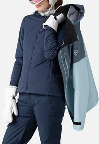 Marineblaue, mit Reißverschluss ausgestattete Jacke mit Kapuze, die ein hellblaues Innenfutter zeigt. Verfügt über weiche Bündchen und ein Logodetail. Das Modell hält eine Jacke und trägt Handschuhe.
