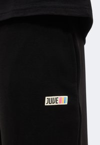 Μαύρο ύφασμα με ένα μικρό ορθογώνιο μπαλώμα με την ετικέτα "JUVE", ακολουθούμενο από τρεις κάθετες ρίγες σε ροζ, κίτρινο και μπλε.