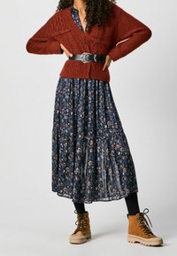 Ein rostfarbener gestrickter Cardigan über einem floralen navy Maxikleid, schwarzer Gürtel mit silberner Schnalle, kombiniert mit hellbraunen Schnürstiefeln.
