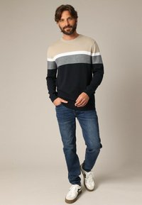 Pull homme à rayures beige, marine et grises ; manches longues, col rond, texture tricotée. Assortie avec un jean bleu et des baskets blanches.