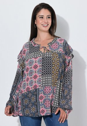 Mujer que lleva una blusa suelta de manga larga con un colorido patrón floral y geométrico en patchwork, sonriendo y mirando hacia un lado.