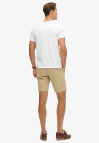 Camiseta blanca de manga corta, pantalones cortos color beige y zapatos de barco rojo oscuro. Los pantalones cortos son de tela caqui con corte recto y bolsillos. Conjunto casual visto desde atrás.