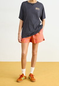 Mörkgrå oversized t-shirt med Roxy-logotyp, rosa texturerade shorts och orange sneaker med gröna ränder, bärs med vita strumpor på en gul golv.