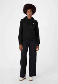 Felpa nera con cappuccio, tasca frontale e coulisse, abbinata a pantaloni scuri larghi e scarpe bianche. Tessuto con texture liscia.