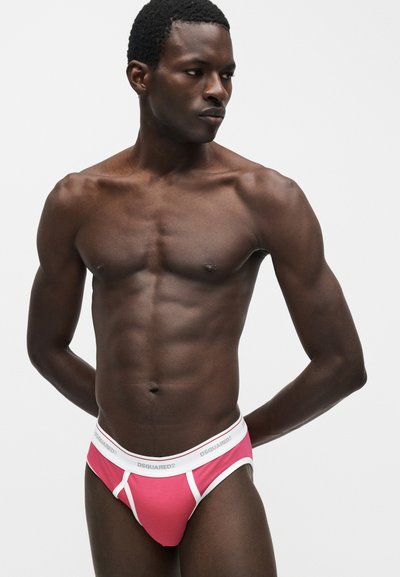 Μυώδης άνδρας με σκούρο δέρμα με ροζ και λευκά εσώρουχα DSQUARED2, στέκεται μπροστά σε απλό ανοιχτό φόντο, κοιτάζοντας προς τα δεξιά.