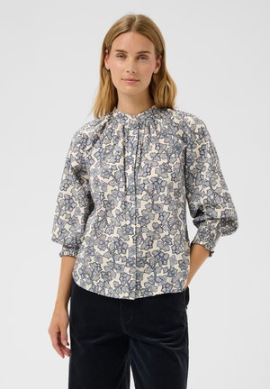 Femme aux cheveux blonds mi-longs portant un chemisier crème à imprimé floral bleu et manches trois-quarts, associé à un pantalon foncé.