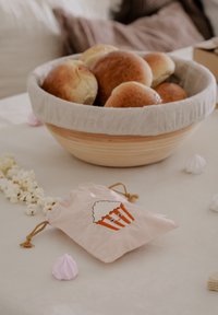 Taška na cukrovú vatu s grafikou popcornu, jemne ružová farba, uzatváranie na sťahovaciu šnúrku. V pozadí je drevená misa s hnedými chlebovými žemľami.