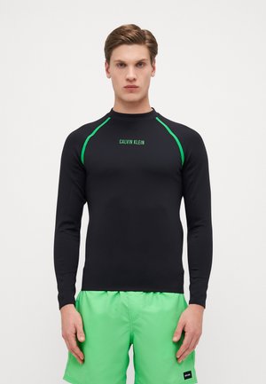 Black long-sleeve rash guard with green accent stitching and "CALVIN KLEIN" logo. Worn with bright green swim shorts, showcasing a fitted design.