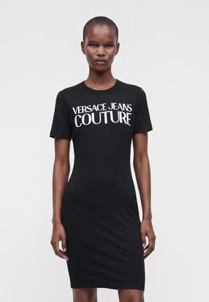 Robe noire cintrée à manches courtes avec texte blanc "Versace Jeans Couture" portée par une femme aux cheveux courts, debout devant un fond uni.