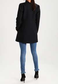 Mujer vistiendo un blazer largo negro, jeans skinny con dobladillo deshilachado azul y botines de tacón alto negros, de pie de espaldas.