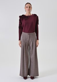 Bordeauxfarbener Pullover mit Rüschenschultern, kombiniert mit weit geschnittenen, karierter Hose mit Knopfverschluss und Falten.