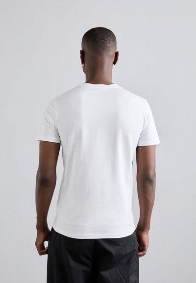 T-shirt en coton blanc à manches courtes, décolleté rond et texture lisse, vue de dos. Un short noir est partiellement visible.