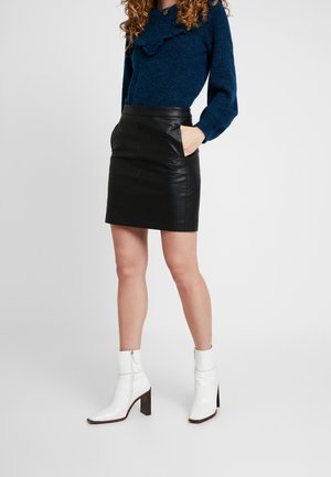 Žena v modrém svetru, černé kožené minisukni s rukama v kapse a bílých kotníkových botách na podpatku stojící na jednobarevném pozadí.