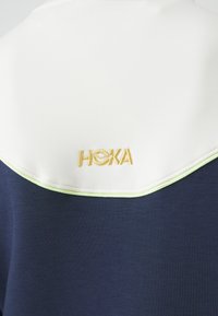 Marineblå treningsgenser med hvit krage med grønn kant, brodert "HOKA"-logo i gull på ryggen, jevn tekstur.