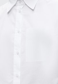 Chemise blanche à boutons avec col et poche poitrine présentant un texte brodé discret.