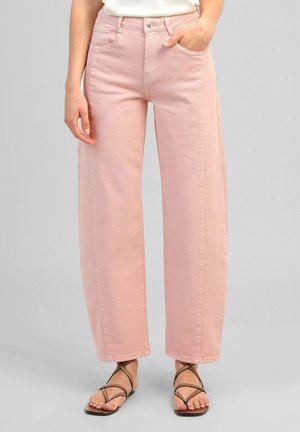 Femme portant un pantalon rose taille haute à jambes droites et des sandales marron à brides, une main dans la poche, sur un fond uni.