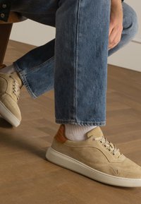 Beige suede sneakers with a rubber sole, featuring a leather accent at the heel and decorative stitching. Worn with light blue denim.