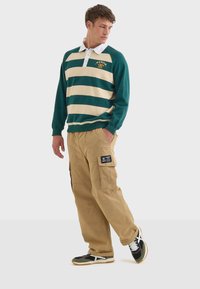 Zeleno-krémový pruhovaný ragbyový dres s bílým límečkem, kombinovaný s khaki cargo kalhotami se bočními kapsami a černým detailem na štítku. Černé a zelené tenisky.