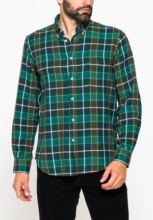 Uomo che indossa una camicia a quadri verde e blu a maniche lunghe e pantaloni neri, in piedi su sfondo bianco.