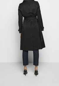 Trench-coat noir à double boutonnage, taille ceinturée et épaulettes, associé à un jean foncé et des chaussures à talons hauts.