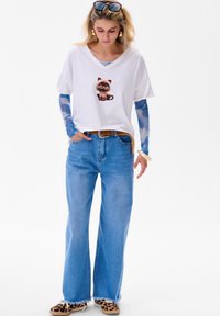 Biała koszulka z krótkim rękawem z grafiką kota, połączona z jasnoniebieskimi szerokimi spodniami dżinsowymi. Dodatki to okulary przeciwsłoneczne i buty w leoparda.