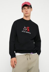 Czarna bluza z haftem przedstawiającym postacie z kreskówek oraz napisem "Forever Hustling". Połączona z beżowymi spodniami z elastycznym paskiem.