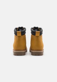Botas de cuero amarillas con cuello acolchado negro, puntera redonda y suela de goma texturizada. Presentan detalles de costura y una lengüeta en la parte trasera.