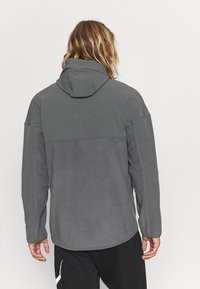 Giacca con cappuccio grigia realizzata in tessuto morbido, caratterizzata da un design a pannelli e dettagli testurizzati sulle maniche, e un orlo arrotondato nella parte inferiore.