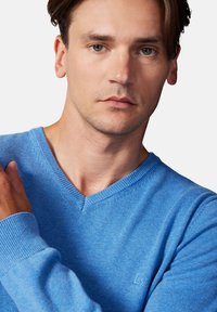 Niebieski sweter w kształcie v z miękkiego materiału dzianinowego, z prążkowanymi mankietami i dołem. Subtelny detal z logo na klatce piersiowej. Gładka faktura.