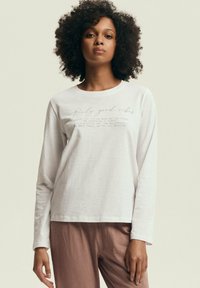 Biała bluzka z długim rękawem wykonana z miękkiego materiału, z prostym okrągłym dekoltem i nadrukowanym jasnobrązowym tekstem z przodu.