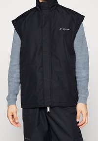 Svart ärmlös väst med hög krage, framdragkedja och sidofickor. Tillverkad av glatt, lätt material. Signaturlogotyp på bröstet.