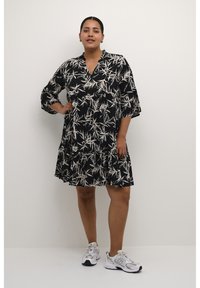 Schwarzes Kleid mit Muster aus weißen Bambusblättern, V-Ausschnitt, drei Viertelärmel und gestuften Rüschensaum, kombiniert mit grauen Sneakers.