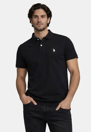 Mann mit kurzen dunklen Haaren, der ein schwarzes Poloshirt mit einem weißen, gestickten Logo trägt und mit den Händen in den Taschen vor einem einfarbigen Hintergrund steht.