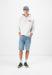 Grå hoodie med logo, blå denimshorts, grønne sneakers og stribede hvide sokker. Tilbehøret inkluderer en blå kasket. Casual outfit design.