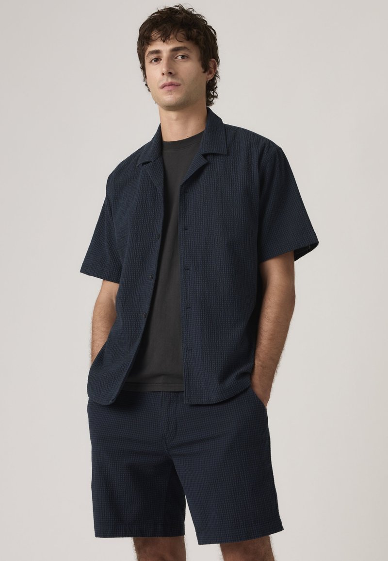 Ung mand i tekstureret mørkeblå kortærmet skjorte og matchende shorts med hænderne i lommerne, stående mod en ensartet lys baggrund.