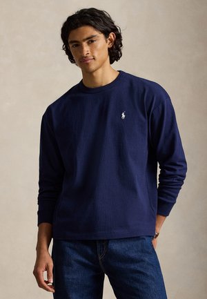 Mladý muž so stredne dlhými tmavými vlasmi, oblečený v námorníckej modrej košeli s dlhým rukávom s malým bielym logom na hrudi a modrými džínami.