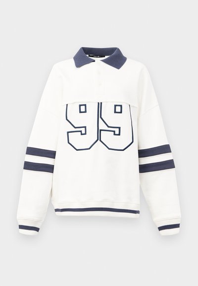 Hvit genser med marineblå krage og detaljer, med nummeret "99" i fet marineblå broderi, og stripete ermer med marineblå bånd.