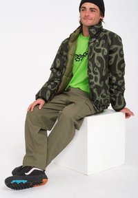 Een gewatteerde jas met een donkergroen abstract patroon, gecombineerd met een felgroen shirt en olijfgroene broek. Zwarte en oranje schoenen met gestructureerde zolen.