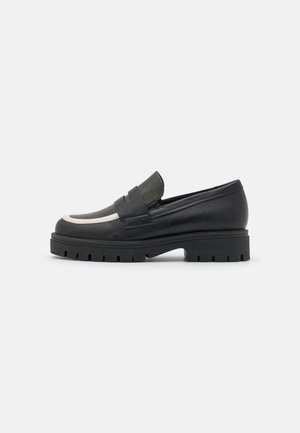Mocassin en cuir noir avec une bande d'accent verte, contrasté par une semelle basse et épaisse avec un motif de crampons prononcé. Détails lisses et texturés.