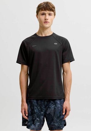 Jeune homme portant un t-shirt de sport noir à manches courtes et un short de sport foncé à motifs, debout devant un fond gris clair.
