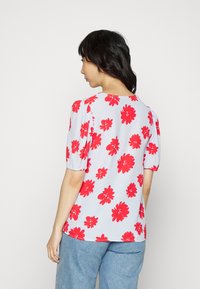 Top de manga curta em tecido azul claro com um padrão floral repetido em vermelho brilhante. Ajuste ligeiramente descontraído com decote redondo.