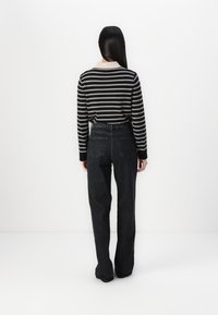 Femme aux longs cheveux raides portant un haut à manches longues rayé noir et blanc avec un col beige et un jean noir taille haute, debout contre un mur blanc.