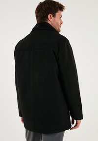 Giacca nera con un design strutturato, caratterizzata da un alto colletto e una texture morbida, esposta da dietro, mettendo in evidenza le sue linee pulite.