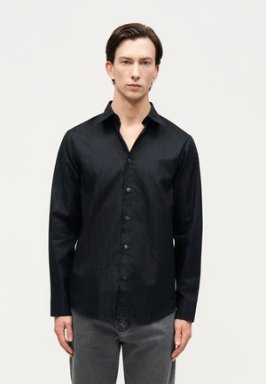 Noor mees, kes kannab musta mustriga pika varrukaga nööbitavat särki ja halli teksapükse, seisab tühja hele tausta ees.