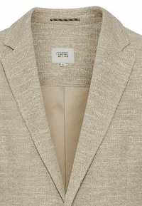 Beige Blazer aus strukturiertem Stoff, mit Schalkragen und einem hellen Futter. Das Innere zeigt ein Netzmuster. Etikett innen sichtbar.